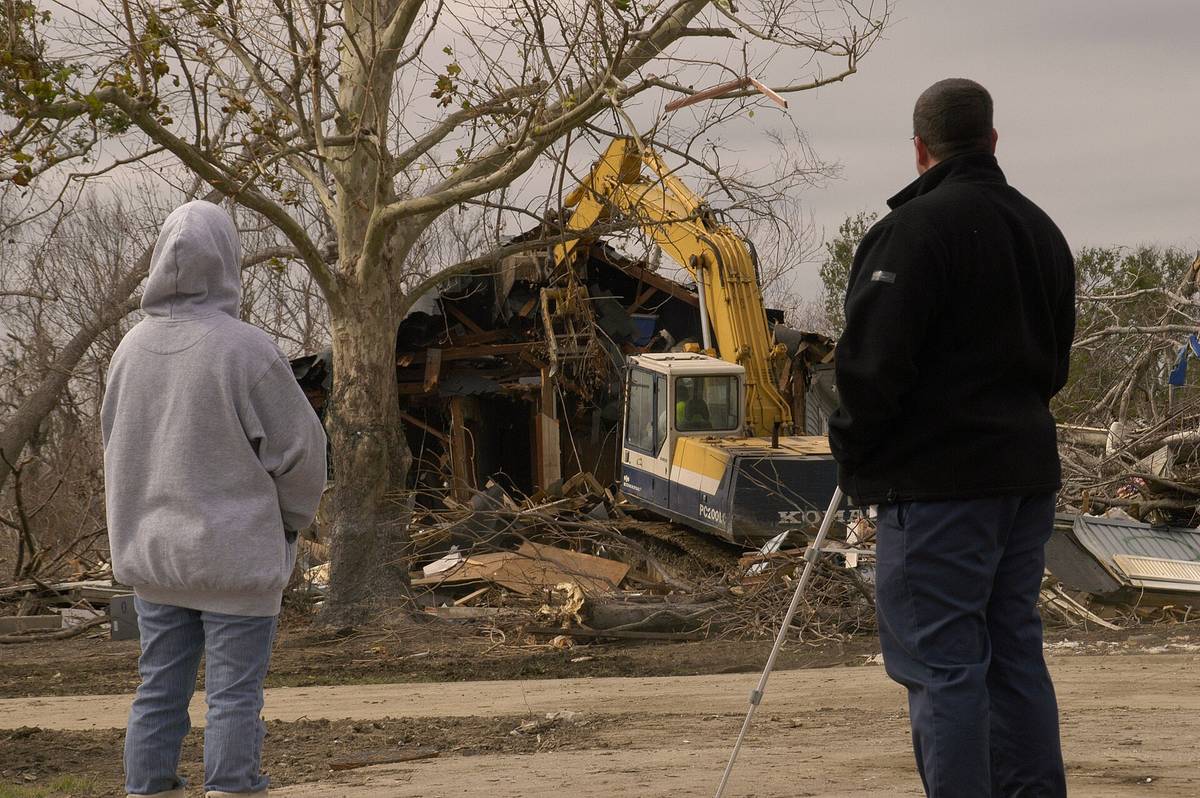 Two people watch an excavator demolish a storm-damaged house