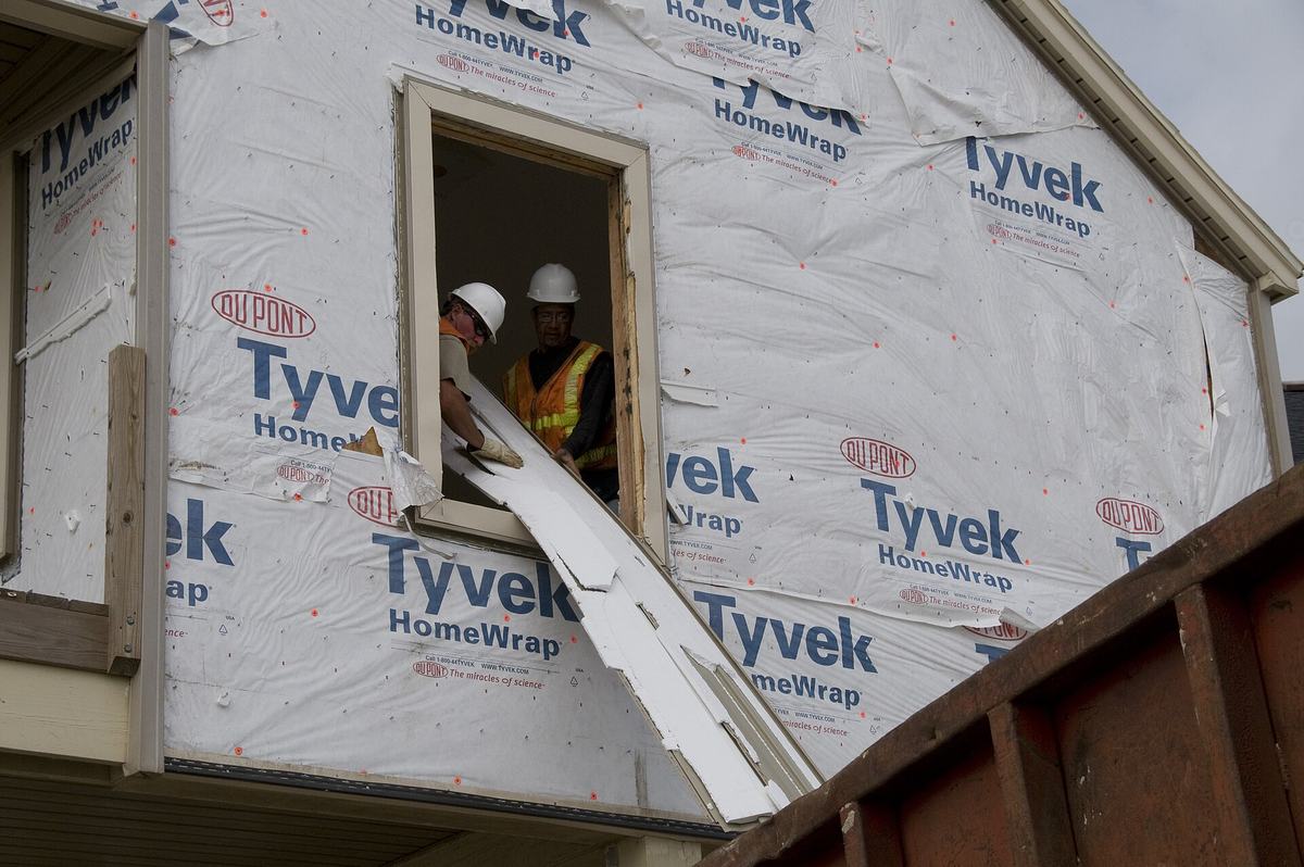 Two workers in hard hats pull material out through a window of a house wrapped for demolition